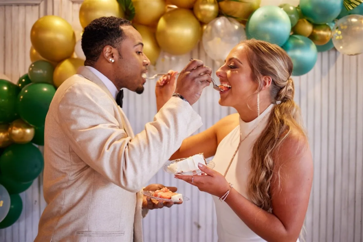 Elegant Nashville wedding cake feeding moment with bride and groom celebrating against gold and green balloon backdrop by A Catered Affair catering