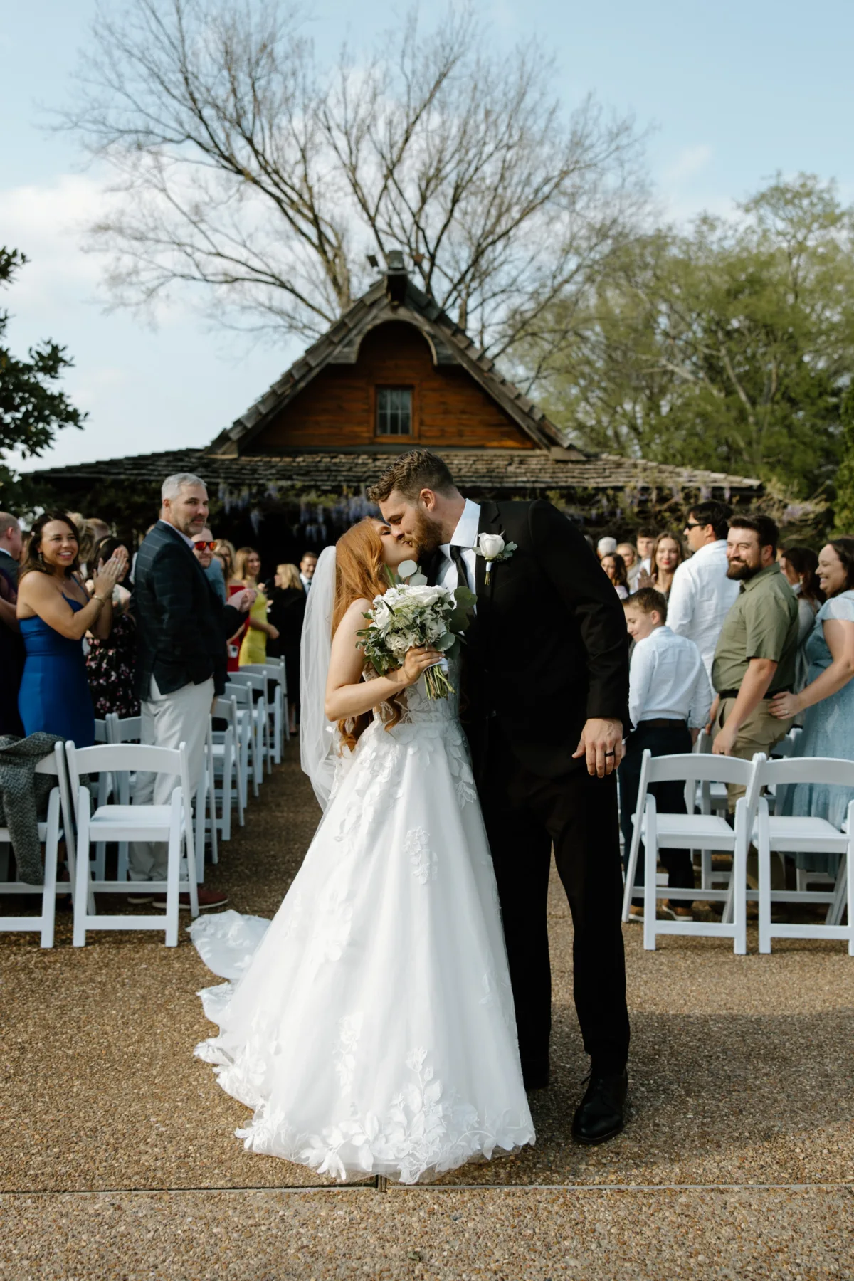 Bride and groom sharing first kiss at outdoor wedding ceremony with rustic wooden chapel and white chairs at Nashville wedding venue